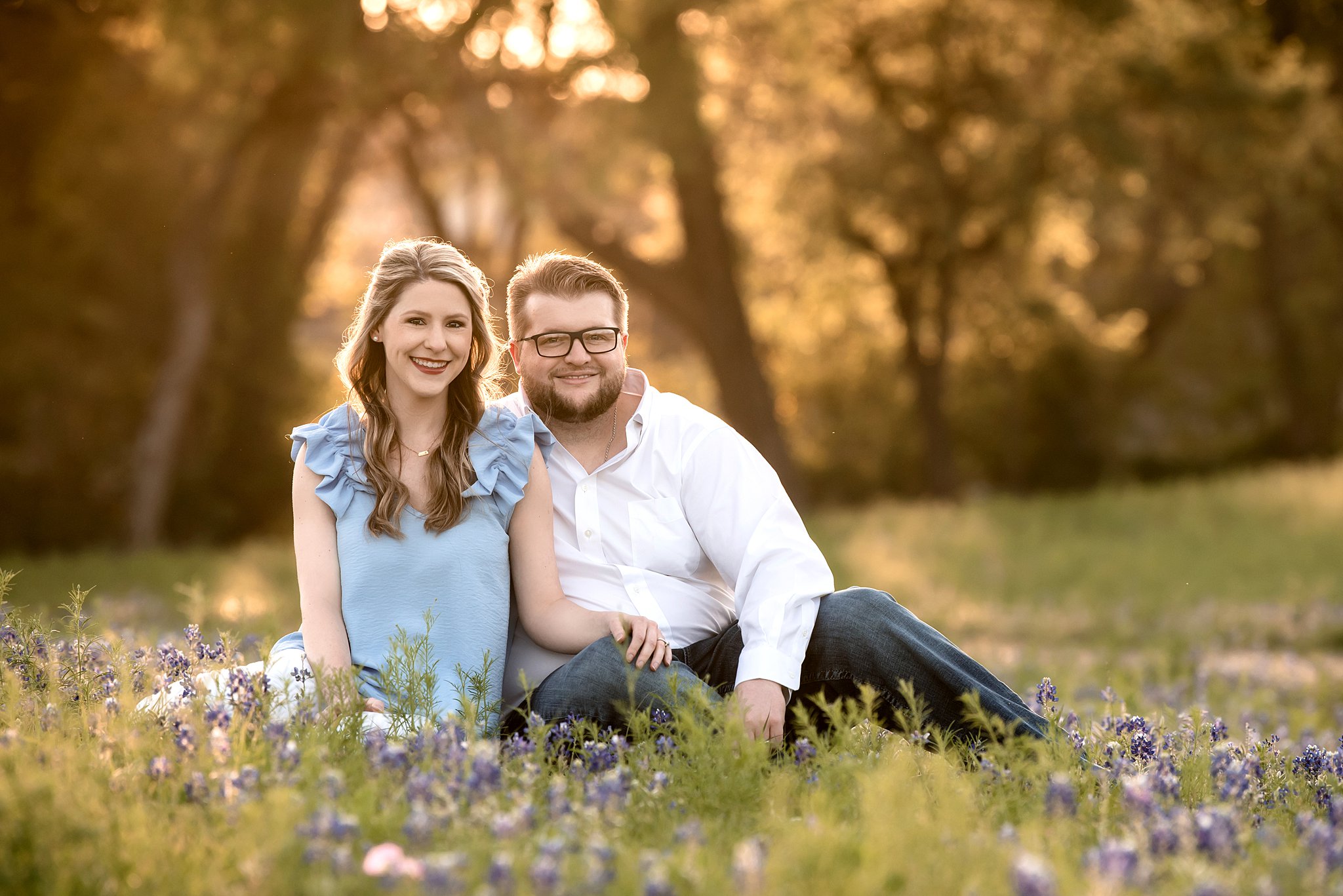 Couple wearing blue and white tops sit in field of purple flowers with sunset coming through the large trees behind them