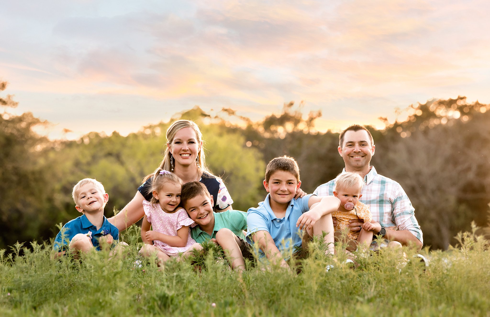 family of seven sits together in a field of green grass at sunset