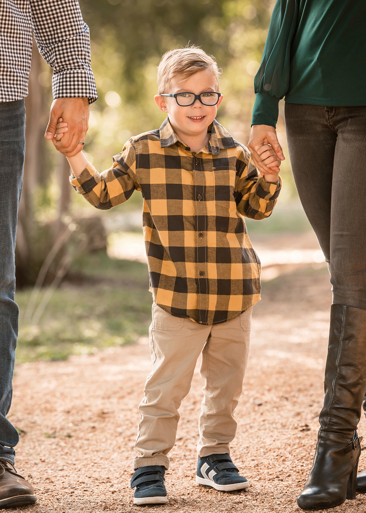 A young boy in a yellow plaid shirt stands in a park path holding his parents' hands on either side