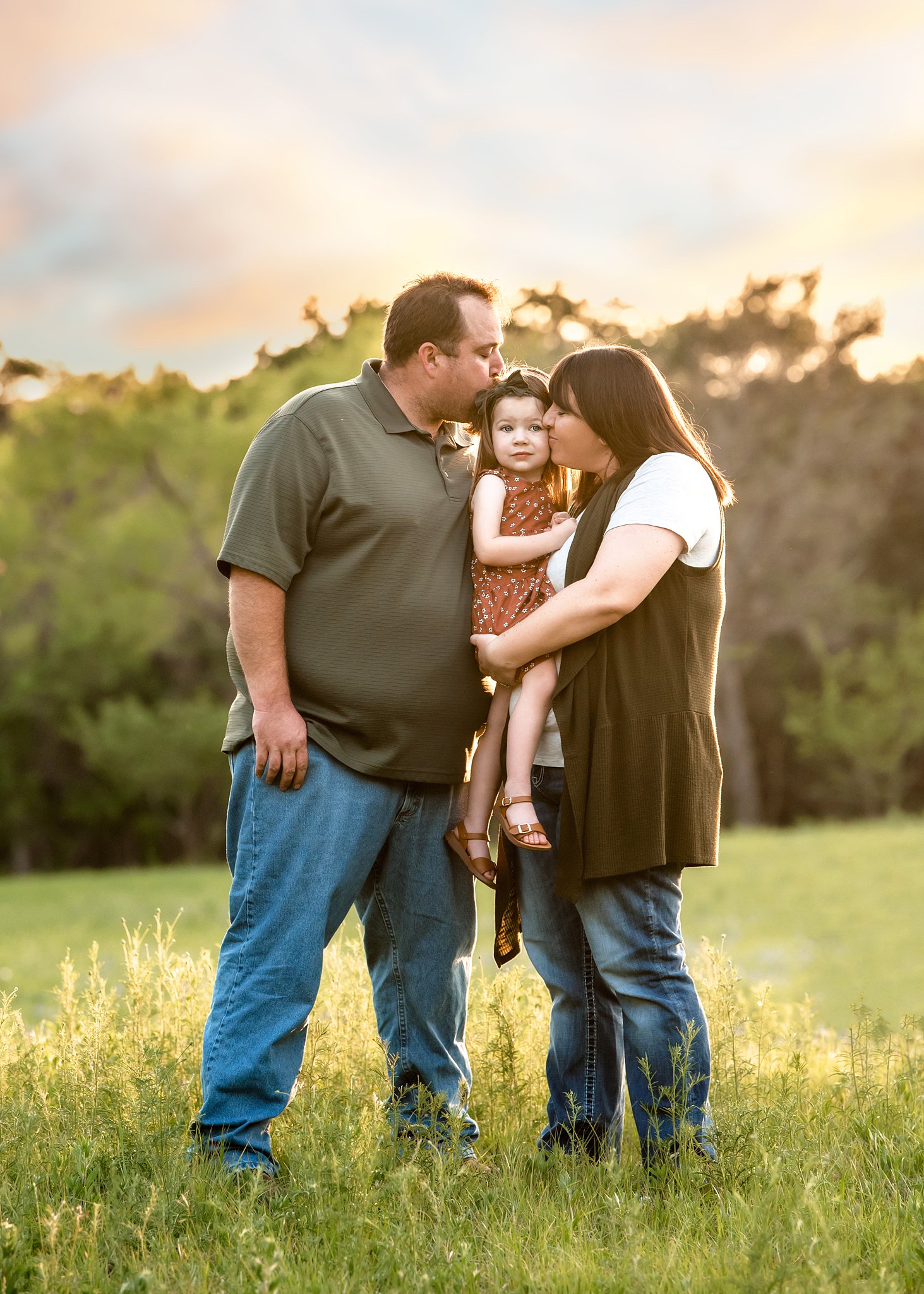 A mother and father stand in a grassy field kissing their young daughter