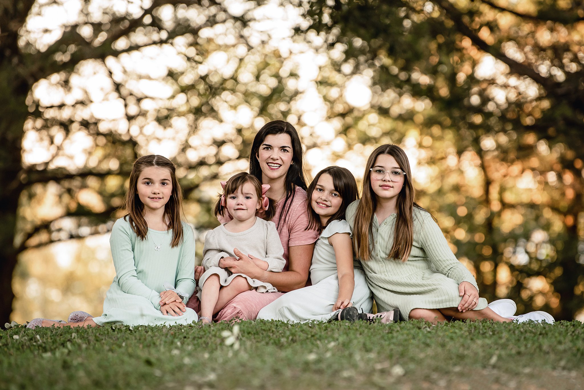A mother sits on the grass of a park at sunset with her four daughters in matching green dresses spas in waco