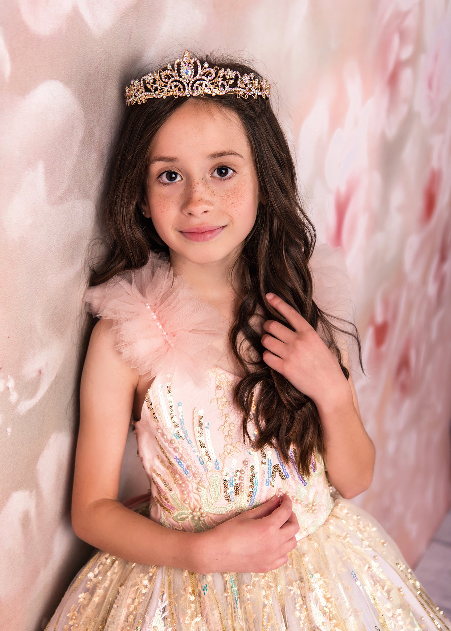 A young girl in a golden princess gown and crown leans against a wall in a studio