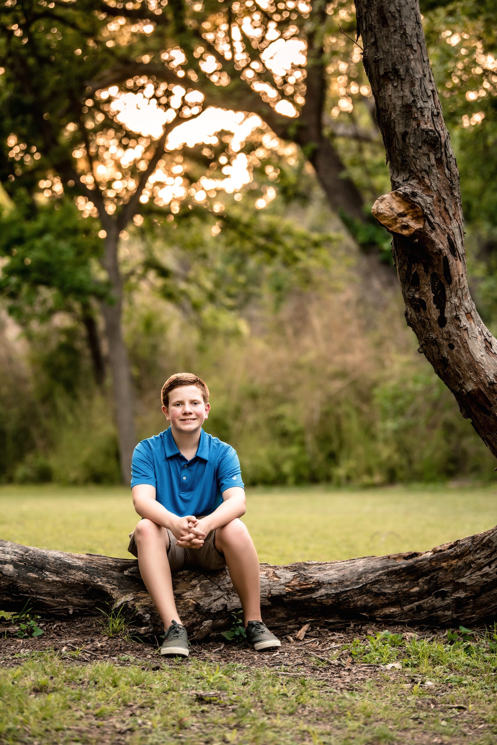 A teenage boy in a blue polo sits on a fallen tree limb in a park at sunset waco public pools