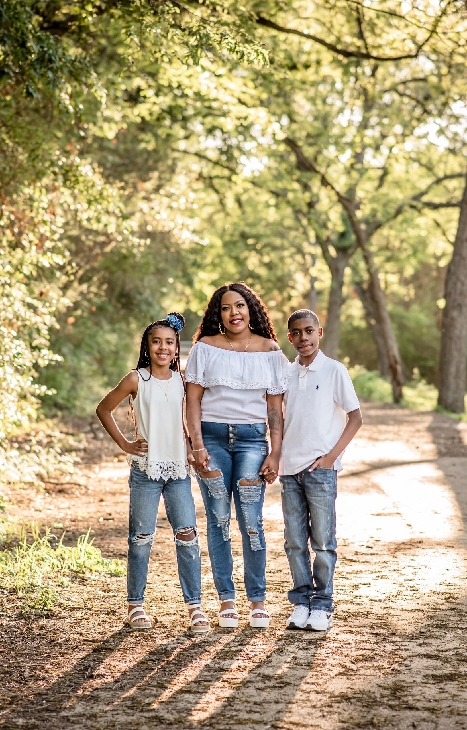A mother in jeans and white top stands in a park path holding hands with her matching son and daughter waco summer activities