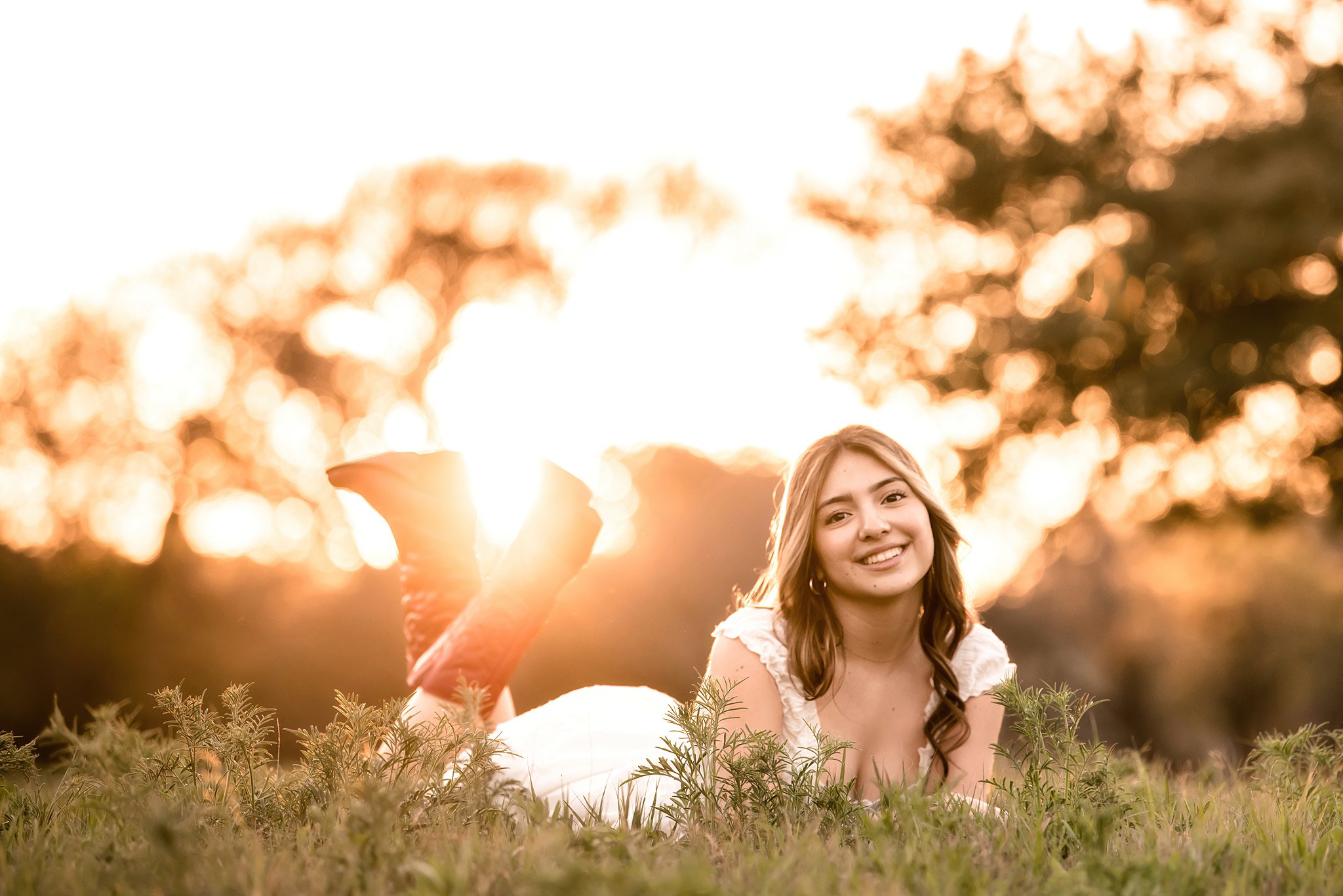 A high school senior lays in a field at sunset in red boots and a white dress