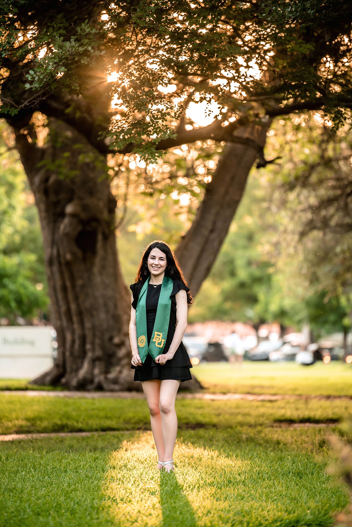 A college graduate stands under a tall tree at sunset displaying her stole
