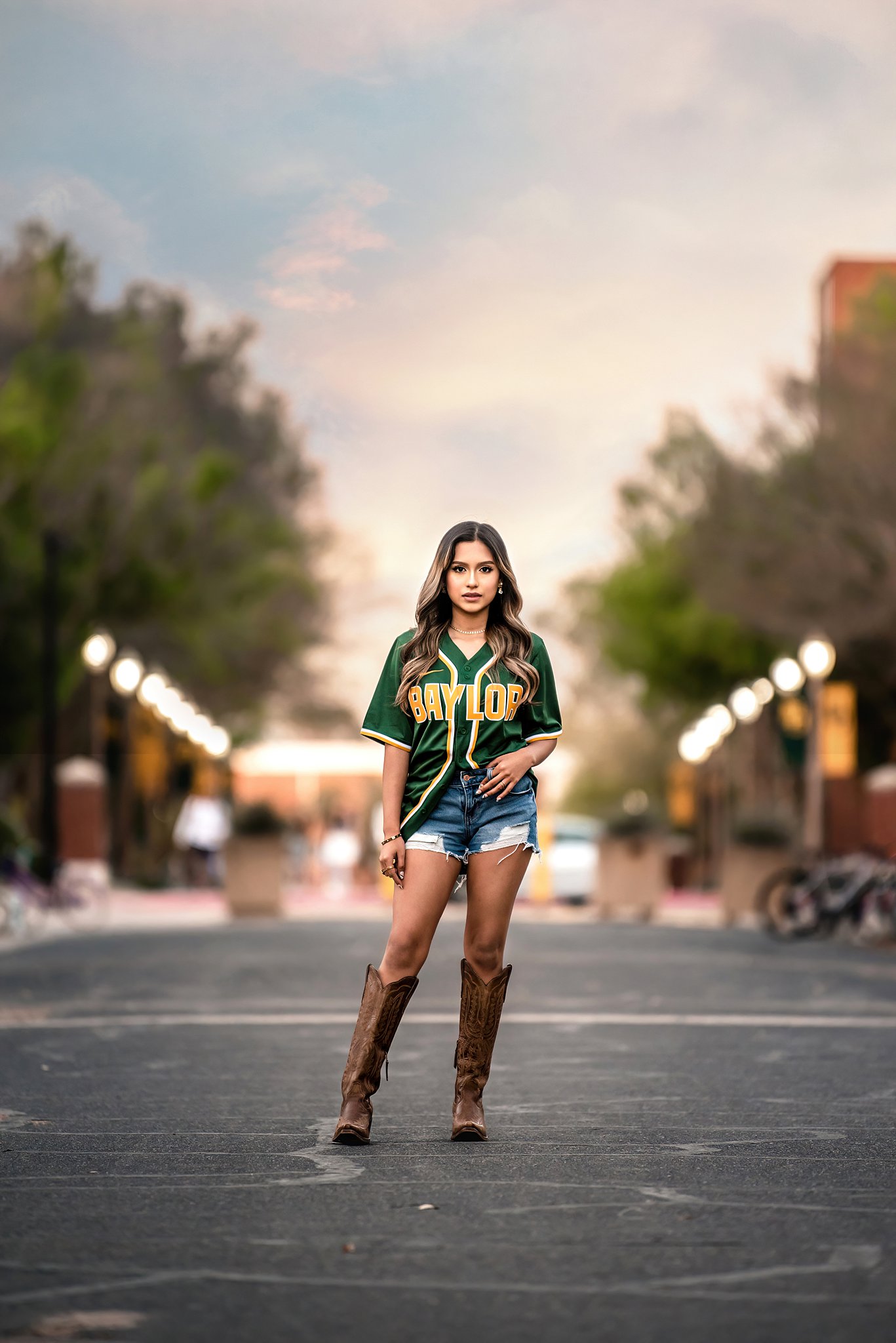 A Baylor graduate stands in a softball jersey and boots in the street at sunset at colleges waco texas