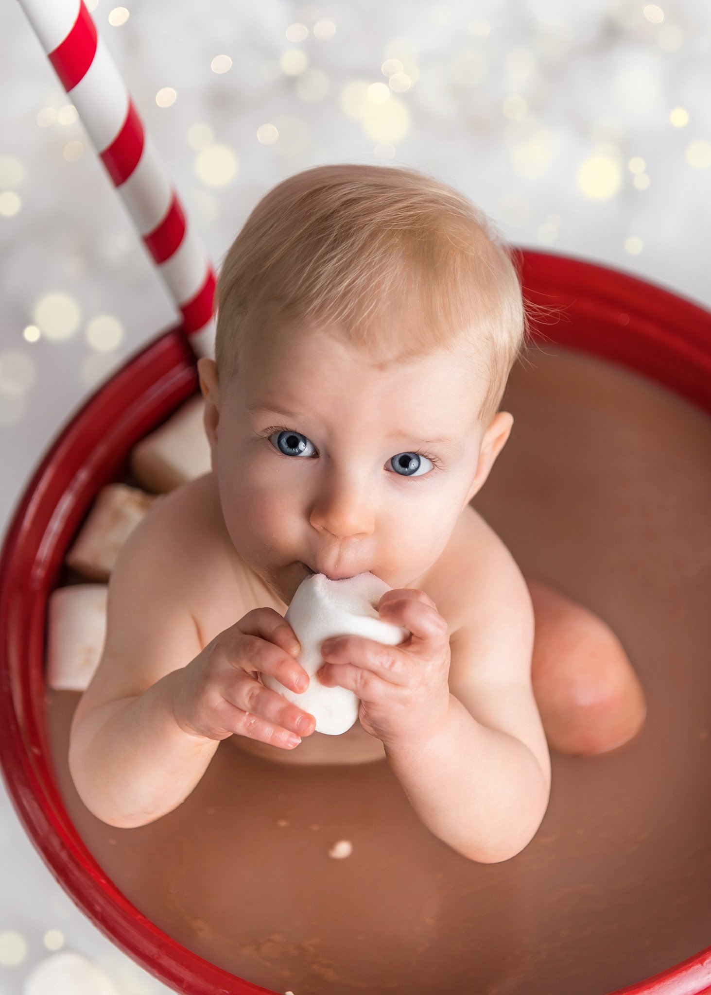 A young toddler sits in a large cup of hot chocolate eating a marshmallow