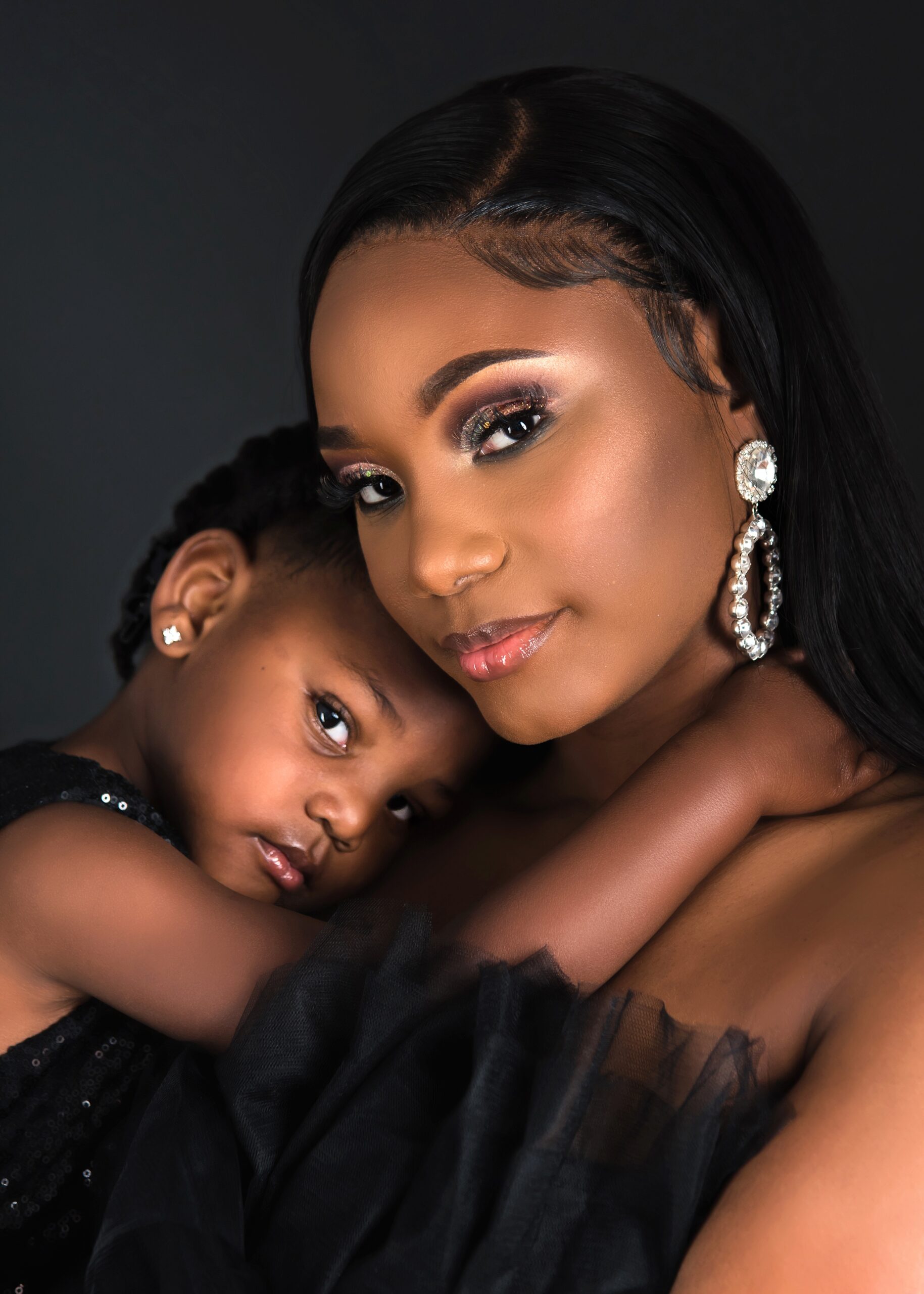A happy mother in a black dress snuggles her toddler daughter in a studio